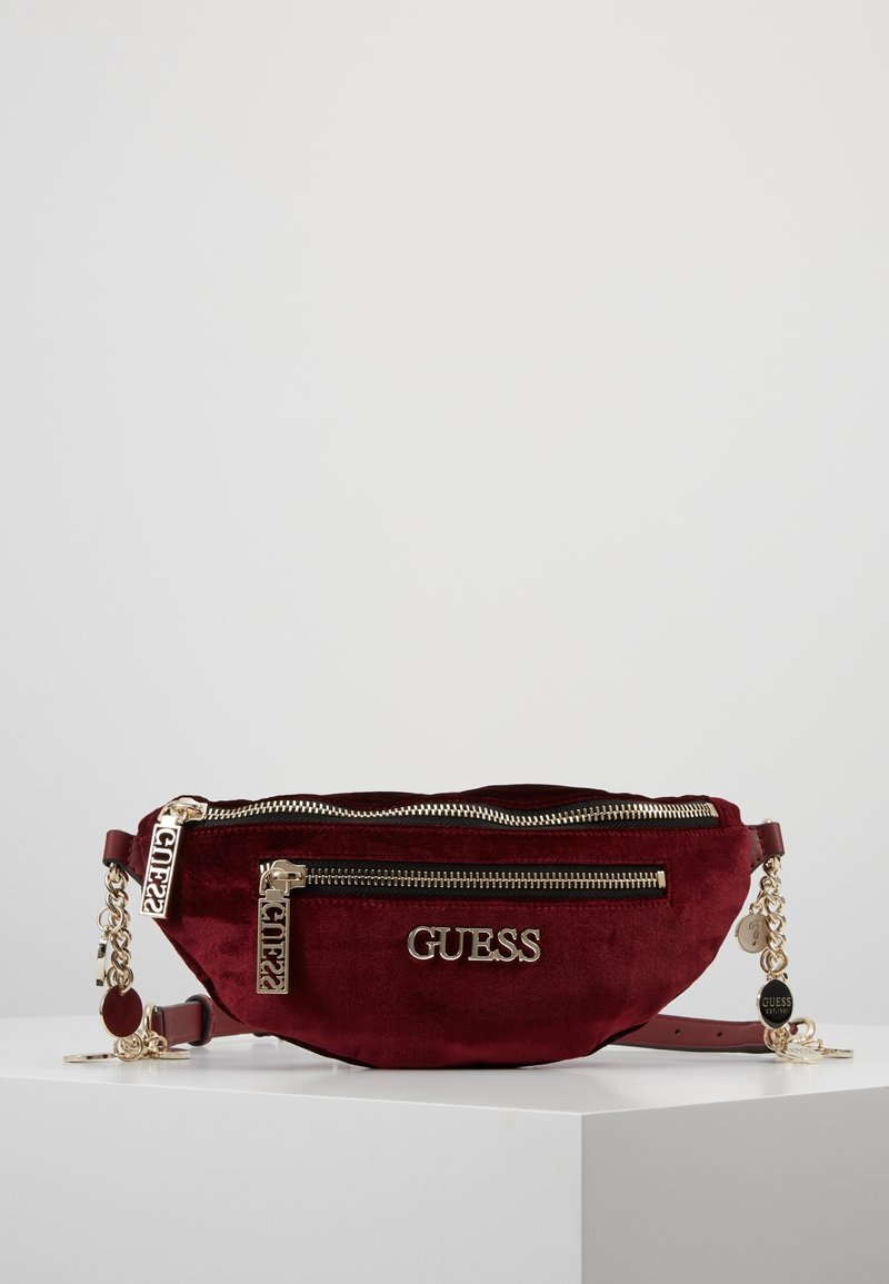 Riñonera de ante burdeos con cremalleras y herrajes en tono dorado, que presenta un logotipo destacado de "GUESS" y detalles de cadena. Dos compartimentos con cremallera.