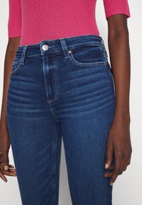 Femme portant un jean bleu foncé et un haut côtelé rose à manches trois quarts, debout devant un fond uni.