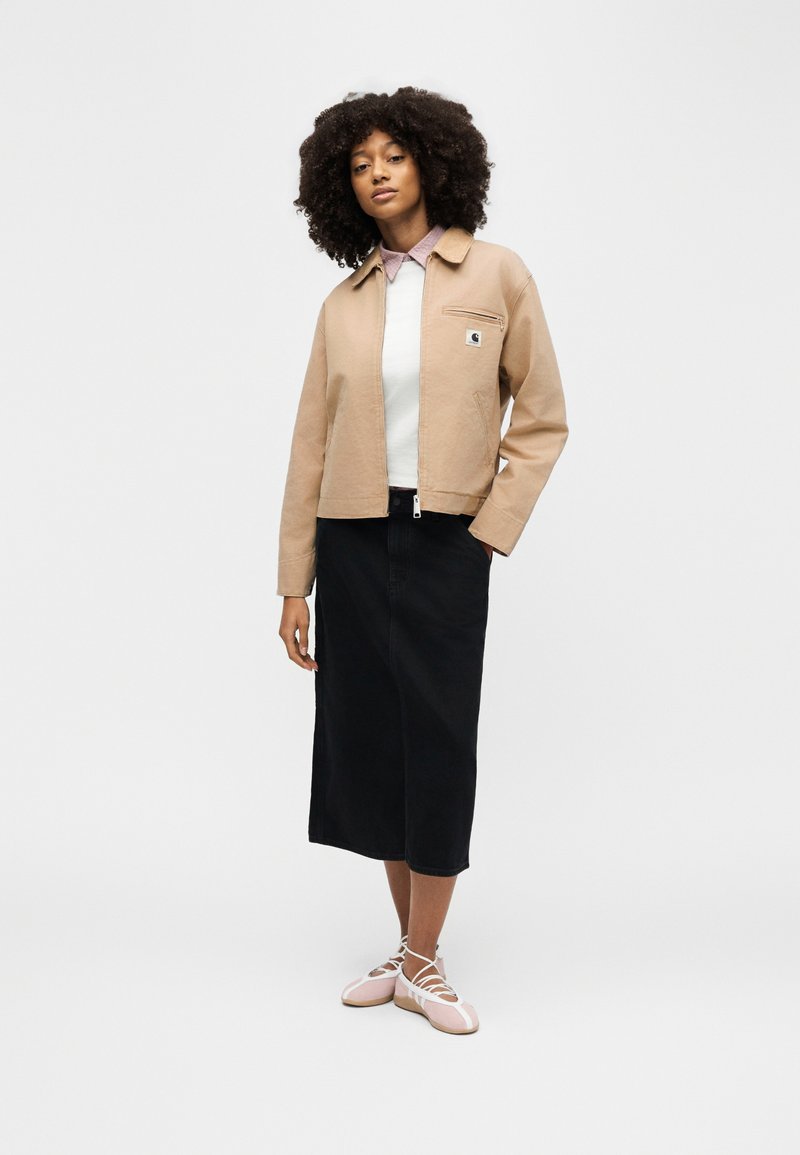 Woman with curly hair wearing a beige zip-up jacket, white top, black midi skirt, and pink ballet flats with lace-up straps, standing.