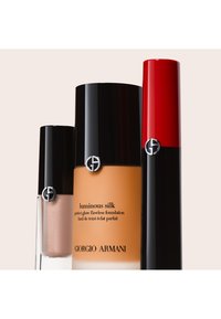 Luminous Silk Foundation in einem bernsteinfarbenen Glas mit schwarzem Deckel; zwei Lippenprodukte in Schwarz und Rot mit silbernen Logo-Akzenten.