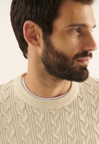 Pulover din tricot cu cabluri, de culoare crem, realizat din fir gros, cu guler rotund și model texturat. Poartă peste o cămașă albă.