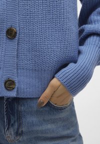 Niebieski sweter na drutach o prążkowanej fakturze, z zapięciem na guziki i luźnym krojem, zestawiony z dżinsami.