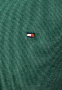 Tissu vert texturé avec un petit logo de drapeau brodé comportant des bandes horizontales blanches, rouges et bleu marine.