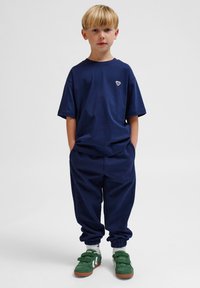 Marinblå bomullströja och matchande joggers, båda med en avslappnad passform. Gröna och vita velcro-sneakers kompletterar outfiten.