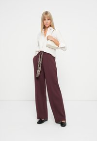 Chemise blanche à manches longues associée à un pantalon large bordeaux avec une ceinture en sash à imprimé léopard. Chaussures plates noires et pochette crème minimaliste.
