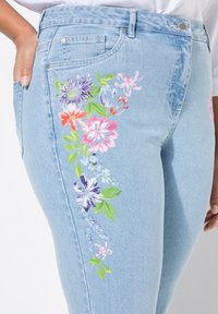 Světle modré džíny vyrobené z denimu, které mají živou květinovou výšivku v růžové, fialové a zelené barvě podél levé nohy, s klasickým designem pěti kapes.
