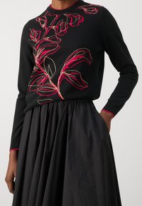Black sweater with red and gold floral embroidery; round neckline and long sleeves; worn with a black, gathered skirt.