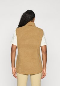Beige fleecevest med høy krage, teksturert stoff og avrundet nederkant, lagd over en lysfarget kortermet skjorte.