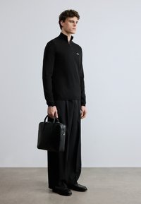 Svart genser med glidelås i halsen, svarte dressbukser og polerte sko. Holder en svart lærmappe med strukturert design og to håndtak.