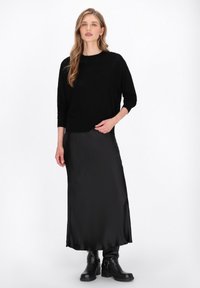Maglione nero a maglia con maniche lunghe, abbinato a una gonna lunga in satin nera e stivali neri alla caviglia, su uno sfondo bianco semplice.