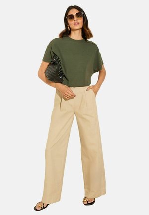 Olivgrünes rippenförmiges Kurzarmshirt, beige weite Hose, schwarze strukturiert Handtasche, übergroße Sonnenbrille und schwarze Sandalen mit goldenen Akzenten.