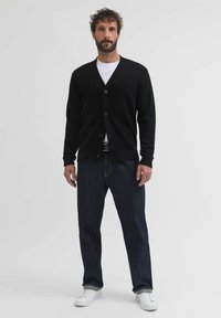 Cardigan negro con escote en V, cinco botones y puños acanalados; usado sobre una camiseta blanca y combinado con jeans oscuros y zapatillas blancas.