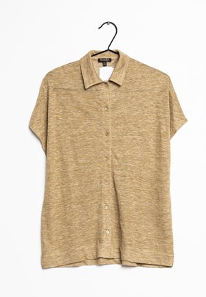 Chemise beige à manches courtes avec col et boutons, suspendue à un cintre noir sur fond blanc.