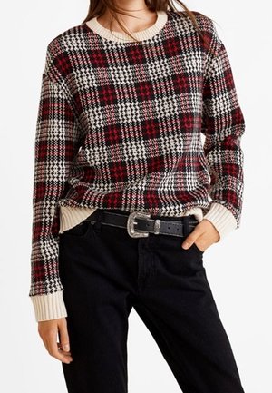 Femme portant un pull en maille à carreaux rouge, noir et blanc avec des poignets et un col crème, rentré dans un pantalon noir avec une ceinture noire.