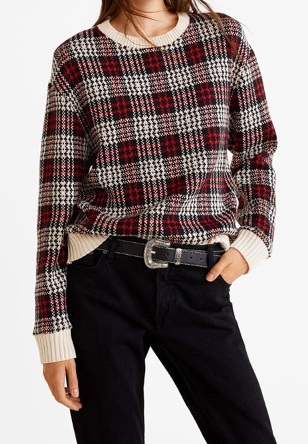 Femme portant un pull en maille à carreaux rouge, noir et blanc avec des poignets et un col crème, rentré dans un pantalon noir avec une ceinture noire.