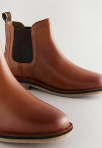 Gli stivali Chelsea in pelle marrone presentano pannelli laterali elasticizzati, un manico per la presa e una suola testurizzata. Il design include cuciture visibili e una finitura liscia.