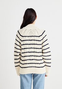 Teksturet kremgenser med marineblå striper, med kabelstrikkedesign og ribbestrikket nederkant. Oversize passform med lange ermer.