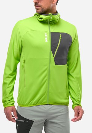 Homme portant une veste zippée vert vif avec une poche poitrine noire et un pantalon gris avec une poche latérale noire, regardant sur le côté.