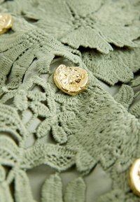 Tissu en dentelle verte avec une broderie florale détaillée et des boutons dorés brillants de forme irrégulière.