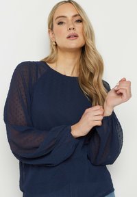 Marinblus med prickig textur, rundad halsringning och ballongärmar. Har genomskinliga ärmar och en lös passform som skapar en elegant silhuett.
