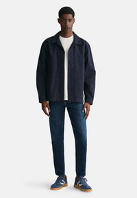 Giacca blu navy in una vestibilità casual con colletto, chiusura a bottoni e tasche anteriori. Abbinata a jeans slim e sneakers blu.