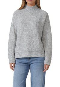 Vrouw in een lichtgrijze gebreide trui met een geribbeld hoge hals, boorden en zoom, gecombineerd met lichtblauwe jeans, staand met ontspannen handen.