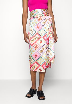 Kobieta ubrana w kolorową spódnicę z motywami zwierzęcymi i roślinnymi, różowy bezrękawnik oraz czarne sandały na obcasie z odkrytymi palcami, stojąca na białym tle.