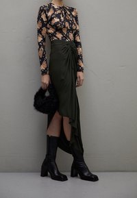 Top con maniche lunghe floreale in nero e beige, abbinato a una gonna drappeggiata verde scuro con uno spacco laterale. Stivali neri fino al ginocchio e una borsa di pelliccia nera.