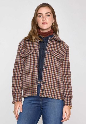 Woman with light brown hair wearing gold hoop earrings, a checked button-up jacket, denim shirt, brown turtleneck, and blue jeans.