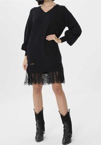 Svart stickad klänning med V-ringning och puffärmar, prydd med en genomskinlig svart spetskant. Kompletterad med spetsdetaljer och en metallbricka.