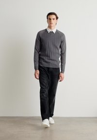 Grauer gerippter Pullover mit einem weißen Hemd darunter, kombiniert mit schwarzen Hosen und weißen Sneakers; steht vor einem neutralen Hintergrund.