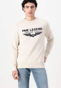 Bež dukserica s okruglim izrezom, dugim rukavima i rebrastim manžetama. Sadrži tamni grafički print s "PME LEGEND" i krilima. Nosena s svijetloplavim trapericama.