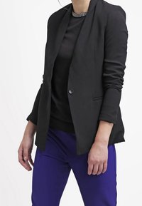 Zwarte blazer met een getailleerd ontwerp, lange mouwen en een enkele knoopsluiting, gecombineerd met een zwart topje en paarse broek. Soepele textuur.