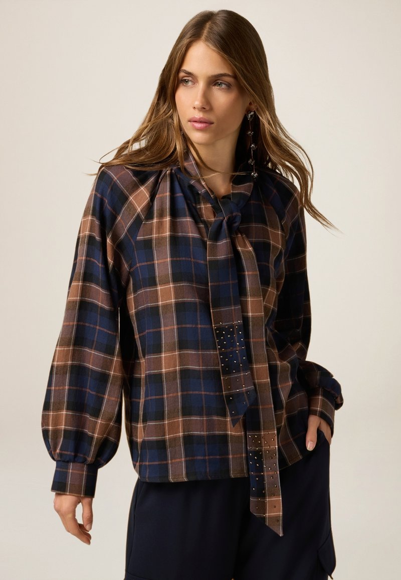 Rutet bluse i mørkeblå, brun og creme med en slipsedetalje ved halsen, lange ballonærmer og nitter foran.