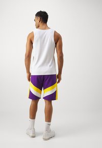 Top sin mangas blanco, pantalones cortos deportivos morados con rayas diagonales amarillas y blancas. Usado con zapatillas blancas y calcetines hasta el tobillo.