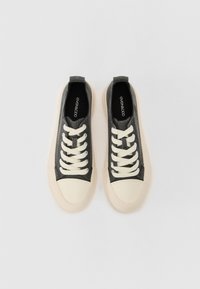 Grå canvas-sneakers med krämfärgade gummisulor och skosnören, rundad tå och texturerad yta. Märkeslogga synlig på innersulorna.