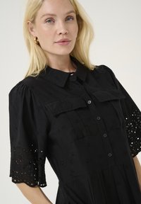 Black button-up shirt dress with short puff sleeves and floral eyelet detailing. Features two chest pockets and a textured fabric.