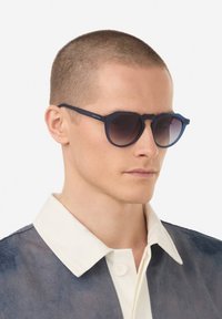 Kulaté sluneční brýle s tmavě modrým matným povrchem, přechodovými čočkami a unikátním mostem. Model nosí košili s límečkem v vrstvených texturách.