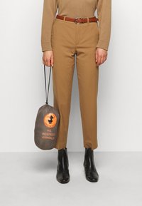 Beige villaneule, ruskeat vyölliset housut, mustat nilkkasaappaat, pitäen harmaata pussukkaa, jossa on oranssi logo, joka sanoo "Kunnioitamme eläimiä."