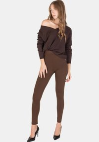 Donna che indossa un maglione marrone scollato sulle spalle, leggings marroni a vita alta e tacchi alti neri, in piedi con una gamba leggermente avanti.