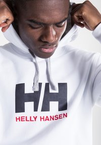Sudadera blanca de mezcla de algodón, con un gran logo negro "HH" y el texto rojo "HELLY HANSEN". Cuerdas de ajuste gris en la capucha.
