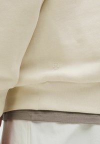 Lys beige sweatshirt med tekstureret strik, der har en ribbet kant og et lille logo forneden. Lagd over en grå skjorte.
