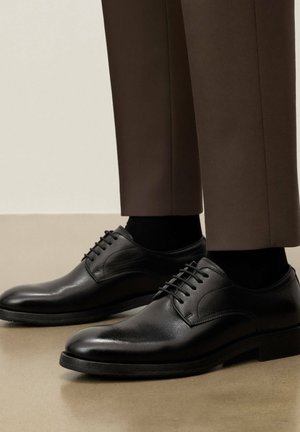 Mustad nahast kingad paeltega, kantud mustade sokkidega ja pruunide rätsepatööna valmistatud pükstega, seistes siledal beežil põrandal.