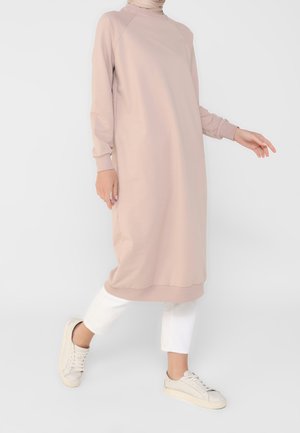 Personne portant une longue robe beige ample par-dessus un pantalon blanc et des baskets beige clair, posant devant un fond clair uni.