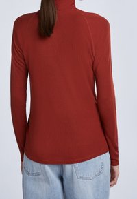Maglione a maniche lunghe color ruggine, con collo alto, vestibilità aderente, realizzato in tessuto elasticizzato, con texture liscia e orlo arrotondato, abbinato a jeans in denim chiaro.