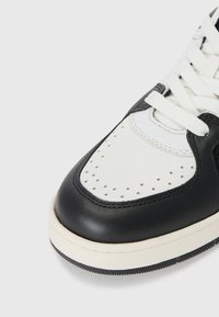 Zwart-witte leren sneaker met geperforeerde neus, contrasterende stiksels, platte witte veters en een gestructureerde rubberen zool.