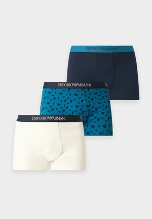 Jeune homme aux cheveux foncés debout pieds nus, portant un boxer blanc Emporio Armani sur un fond clair uni.