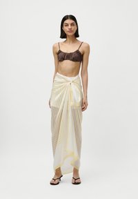 Haut de bikini marron à volants avec fines bretelles, associé à un paréo transparent crème orné de rayures géométriques jaunes, noué à la taille. Tongs noires.