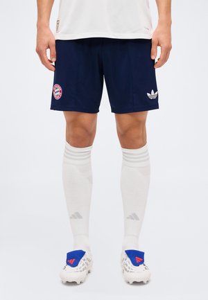 Męskie nogi w granatowych spodenkach Adidas z logo Bayernu Monachium, białe paskowane podkolanówki oraz białe korki piłkarskie z niebieskimi akcentami na pięcie.