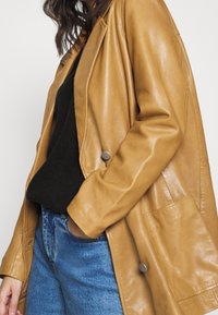 Tan läderjacka med en slät textur, designad med en lös passform, stora fickor och silverfärgade knappar. Tillsammans med en svart tröja och blå jeans.